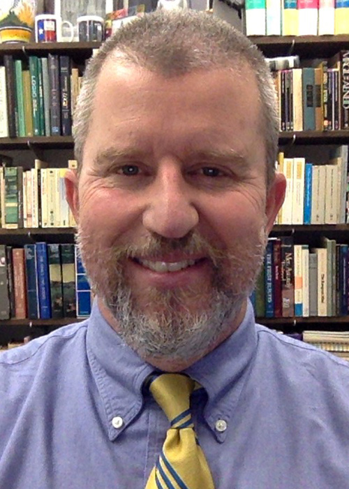 Headshot of man with light hair wearing light shirt and tie against background with bookshelves.